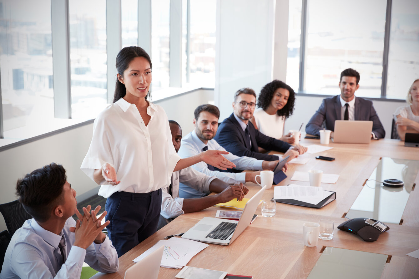 Businesswoman Stands To Address Meeting Around Board Table Businesswoman Stands To Address Meeting Around Board Table